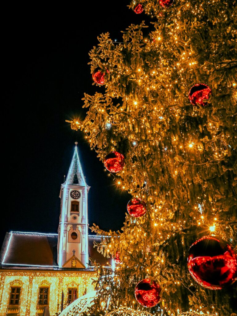 Christmas tree decorated with red baubles and warm lights beside an illuminated church bell tower at night during Advent