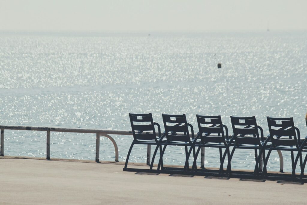 A row of empty chairs on a waterfront facing a glittering sea in hard midday light