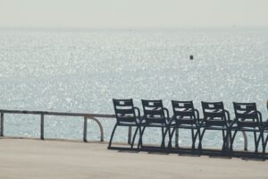 A row of empty chairs on a waterfront facing a glittering sea in hard midday light